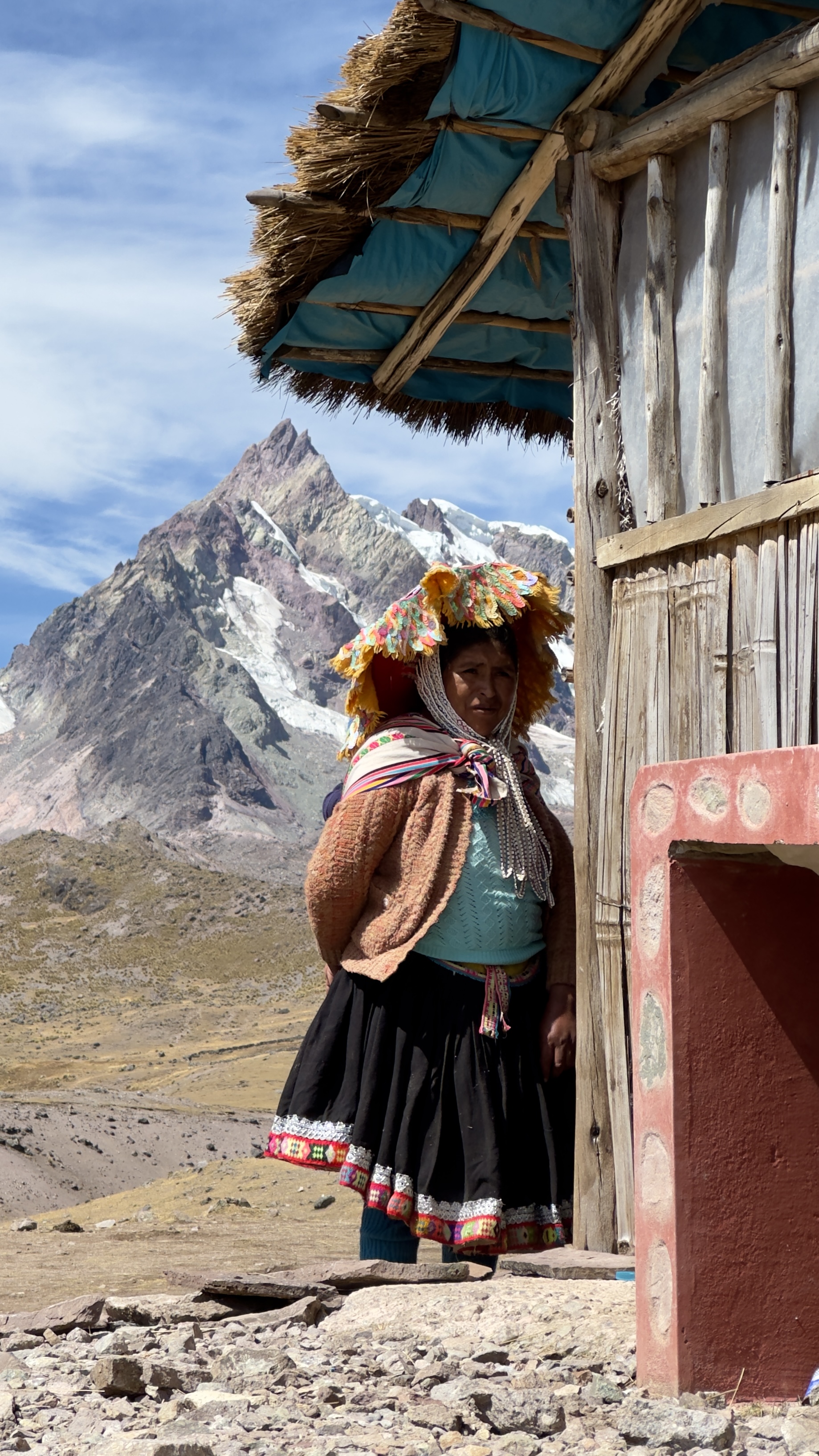 Indigenous woman in traditional colorful dress standing outside mountain hut with peaks behind