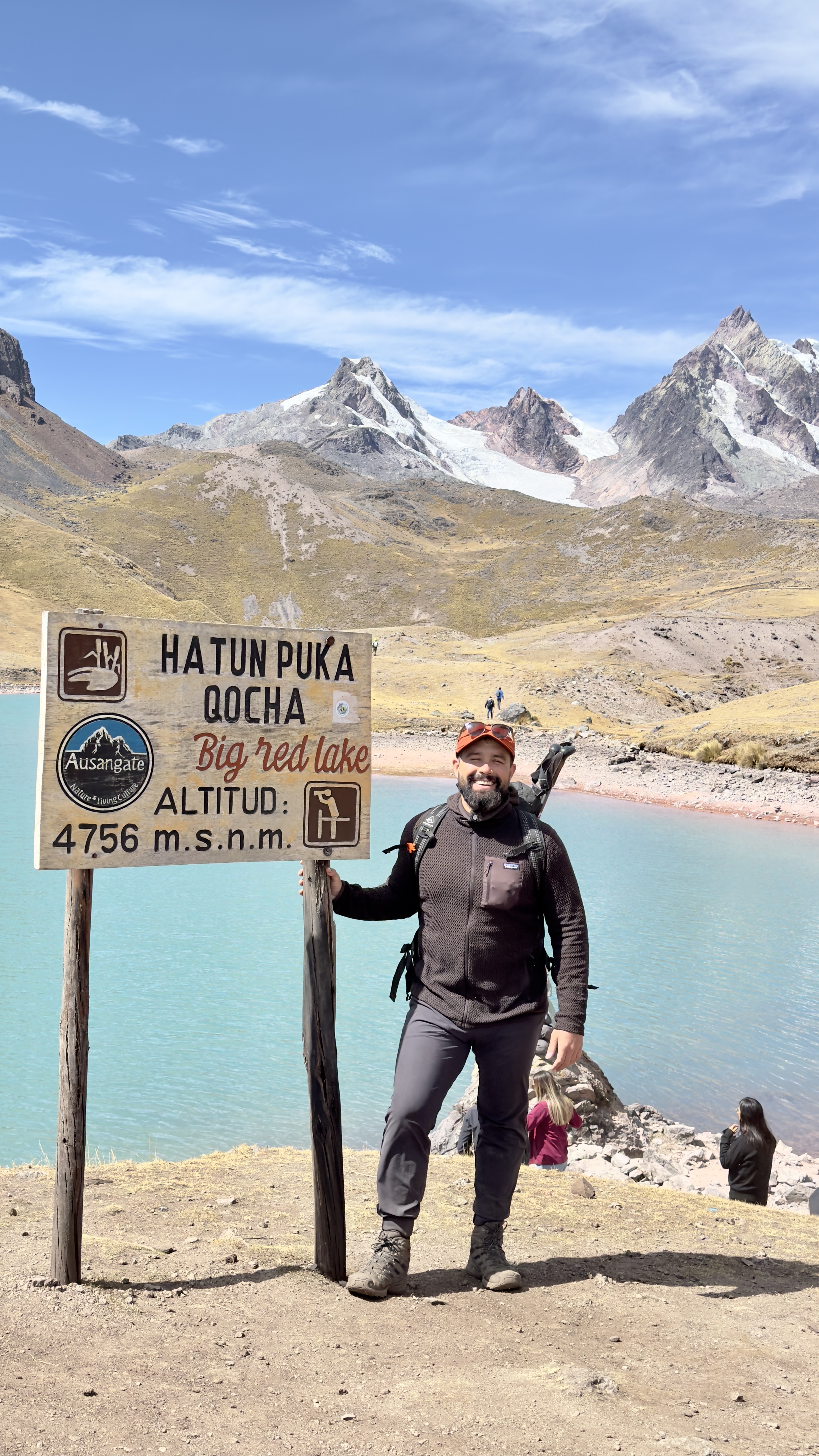 Hiker standing triumphantly by turquoise high-altitude lake sign with snow-capped peaks behind